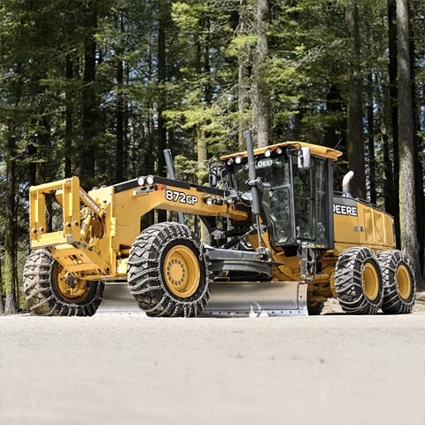 A John Deere 872 GP Motor Grader parked in a forested area, equipped with snow chains on its tires.